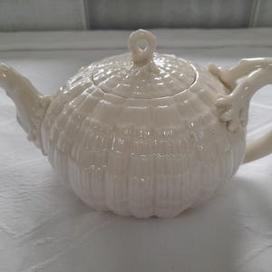 以下が含まれることがあります： 貝殻のような質感の白い陶器製のティーポット。蓋と、巻かれた葉の形をした取っ手があります。