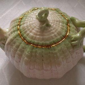 Puede incluir: Una tetera blanca con una tapa verde y dorada y un asa de rana. La tetera está decorada con un patrón floral.
