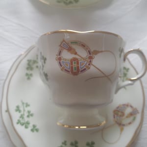 Pode incluir: Conjunto de chá creme com acabamento dourado e design de nó celta. A chávena e o pires apresentam um padrão de trevos verdes. A chávena tem uma base de pedestal e uma pega curva.