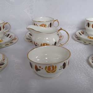 Può includere: Un servizio da tè vintage con tazze, piattini e piatti in porcellana bianca con bordi dorati. Il set include una lattiera e una zuccheriera. Ogni pezzo è decorato con un motivo di foglie marroni, arancioni e verdi e un emblema dorato.