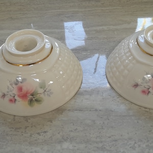 Puede incluir: Dos candelabros de cerámica blanca con borde dorado y diseño floral. Cada candelabro tiene una pequeña asa y una abertura central para una vela.