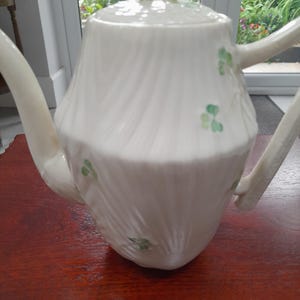 Op de afbeelding: Witte keramische theepot met deksel en handvat. De theepot heeft een textuur en is versierd met groene klaverontwerpen. Het deksel heeft een ringvormige handgreep met groene en gele accenten. De theepot staat op een donker houten oppervlak.