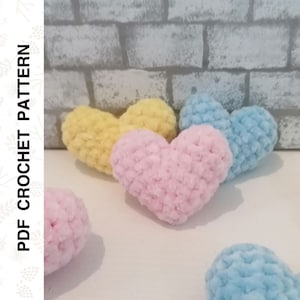 May include: Four crocheted hearts in pink, yellow, and blue. The hearts are made with a fuzzy yarn and are sitting on a white surface.