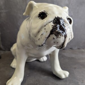 May include: A white ceramic bulldog ornament with black markings on its face. The bulldog is depicted in a sitting pose, with a glossy finish. The figurine has detailed features and is a decorative item.