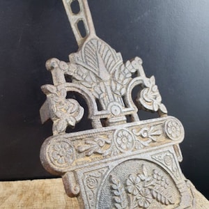 May include: A cast iron fireplace grate with an ornate design featuring floral motifs and a decorative handle. The grate is in a rustic brown colour and has a worn, antique look.