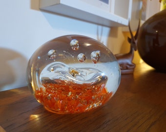 May include: A clear glass paperweight with embedded orange and gold accents. The paperweight has a rounded shape with air bubbles suspended within the glass. It sits on a wooden surface.
