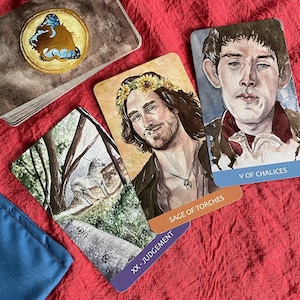 May include: Three tarot cards on a red fabric background. The cards are "Sage of Torches", "V of Chalices", and "XX - Judgement". The card "Sage of Torches" features a man with a sunflower crown and a necklace. The card "V of Chalices" features a man with dark hair and a white bird. The card "XX - Judgement" features a white unicorn in a forest.