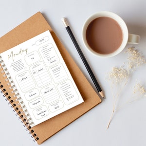 May include: A white planner with a gold inscription that reads "Monday" and a section for daily tasks, appointments, and meal planning. The planner is open on a brown notebook with a black pencil and a cup of coffee on a white surface.