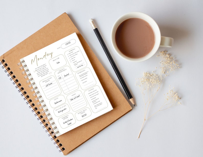 May include: A white planner with a gold inscription that reads "Monday" and a section for daily tasks, appointments, and meal planning. The planner is open on a brown notebook with a black pencil and a cup of coffee on a white surface.