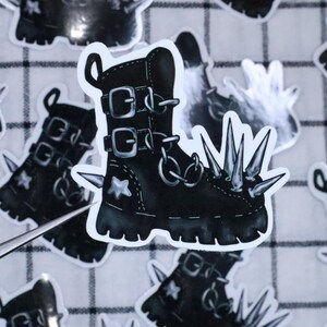 May include: Black and white sticker of a punk rock boot with spikes. The boot has buckles and chains.