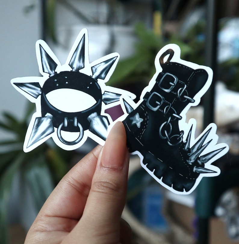 May include: Two black and white stickers. One sticker is a spiked collar with a silver buckle. The other sticker is a black boot with silver spikes and buckles.