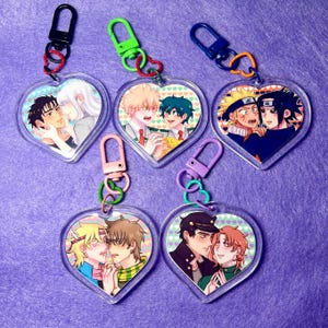 May include: A collection of heart-shaped keychains featuring anime-style illustrations of couples. Each keychain has a clear acrylic base with a colorful image and a metal clasp. The keychains are attached to a purple surface.