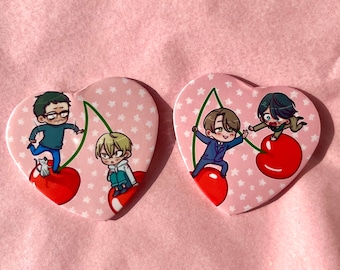 May include: Two heart-shaped pins with a pink background and white stars. Each pin features a cartoon illustration of characters on top of cherries. One pin features a man with glasses and a cat, and the other pin features two men.