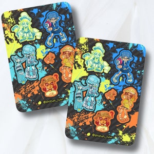 Puede incluir: Dos hojas de pegatinas coloridas con una variedad de personajes de dibujos animados. Los personajes llevan todos atuendos diferentes y tienen diferentes expresiones. Las pegatinas están sobre un fondo negro con salpicaduras de pintura naranja, amarilla y azul. El texto "@shizun_luvr" está en la parte inferior de cada hoja.