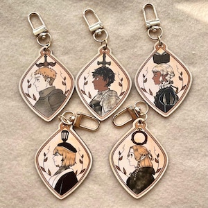 Può includere: Cinque portachiavi in legno con chiusure color argento. Ogni portachiavi presenta un design a forma di diamante con un ritratto di profilo, circondato da un bordo decorativo. I ritratti raffigurano vari personaggi con diverse acconciature e abiti.