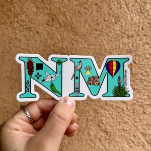 May include: A turquoise blue sticker with the letters NM in white. The letters are filled with various icons representing New Mexico, including a chili pepper, a skull, a roadrunner, a UFO, a hot air balloon, a ski lift, a pueblo, and a yucca plant.