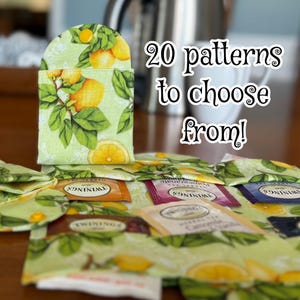 May include: A light green tea bag holder with a lemon and leaf pattern, holding tea bags. The text "20 patterns to choose from!" is displayed in white script. A yellow button secures the holder.