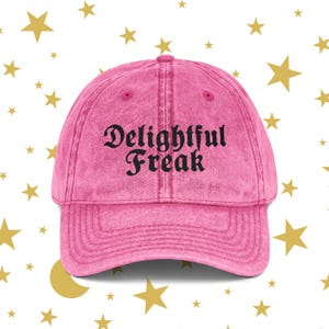 Puede incluir: Gorra de béisbol rosa con las palabras "Delightful Freak" en letras góticas negras. La gorra tiene una visera curva y un aspecto vintage descolorido. El fondo presenta estrellas doradas.