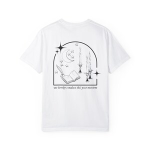 May include: White t-shirt with a black graphic design on the back. The design features a crescent moon, stars, candles, an open book, and the text "we hereby conduct this post mortem."