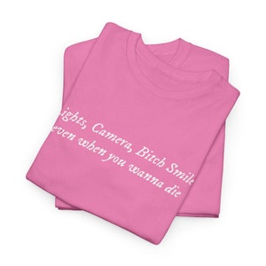 May include: Two folded pink t-shirts with white text that reads "Lights, Camera, Bitch Smile even when you wanna die."