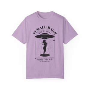 Könnte beinhalten: Ein lavendelfarbenes T-Shirt mit einem schwarzen Grafikdesign, das eine Frau zeigt, die unter einer fliegenden Untertasse steht. Der Text "Female Rage The Musical" ist in einem Kreis um die Untertasse geschrieben. Der Text "Starring Taylor Swift as the tortured poet" steht unter der Grafik.