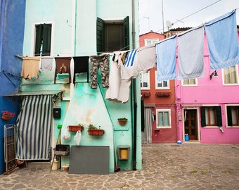 Può includere: Un vicolo stretto in una città europea con edifici colorati. I vestiti sono appesi a uno stendino teso tra gli edifici. Gli edifici sono dipinti in tonalità di blu, verde, rosa e arancio.