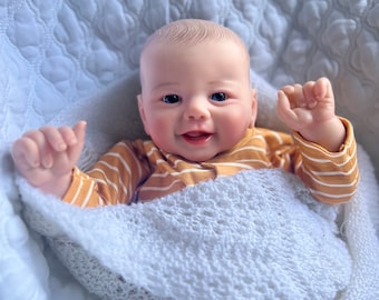 May include: A smiling baby doll with blue eyes and a light brown complexion is wearing a yellow and white striped shirt and is wrapped in a white crocheted blanket.