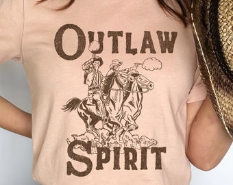 Puede incluir: Una camiseta rosa con un gráfico marrón de un vaquero montando un caballo. El gráfico dice "OUTLAW SPIRIT".