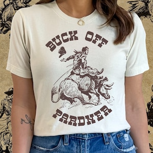 May include: A woman wearing a white t-shirt with a brown graphic of a cowboy riding a bull. The graphic says "Buck Off Pardner".