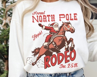 Può includere: Felpa bianca con un'illustrazione rossa e marrone di Babbo Natale a cavallo. Il testo "Annual North Pole Rodeo Dec. 25th" è stampato sulla felpa.