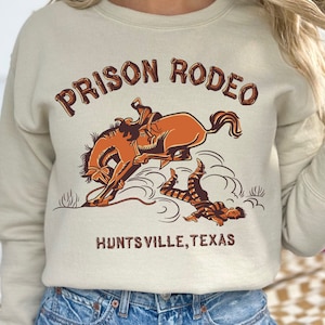 Puede incluir: Una sudadera beige con un estampado gráfico de una persona montando un caballo que se encabrita. El texto "Prison Rodeo" está impreso en marrón y naranja por encima del gráfico. El texto "Huntsville, Texas" está impreso en marrón por debajo del gráfico.