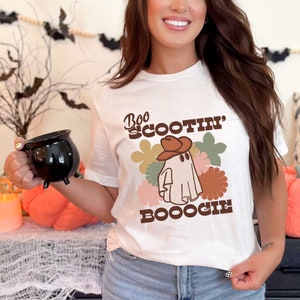 May include: White t-shirt with a brown and white ghost wearing a cowboy hat. The text "Boo Scootin' Booogie" is printed on the shirt.
