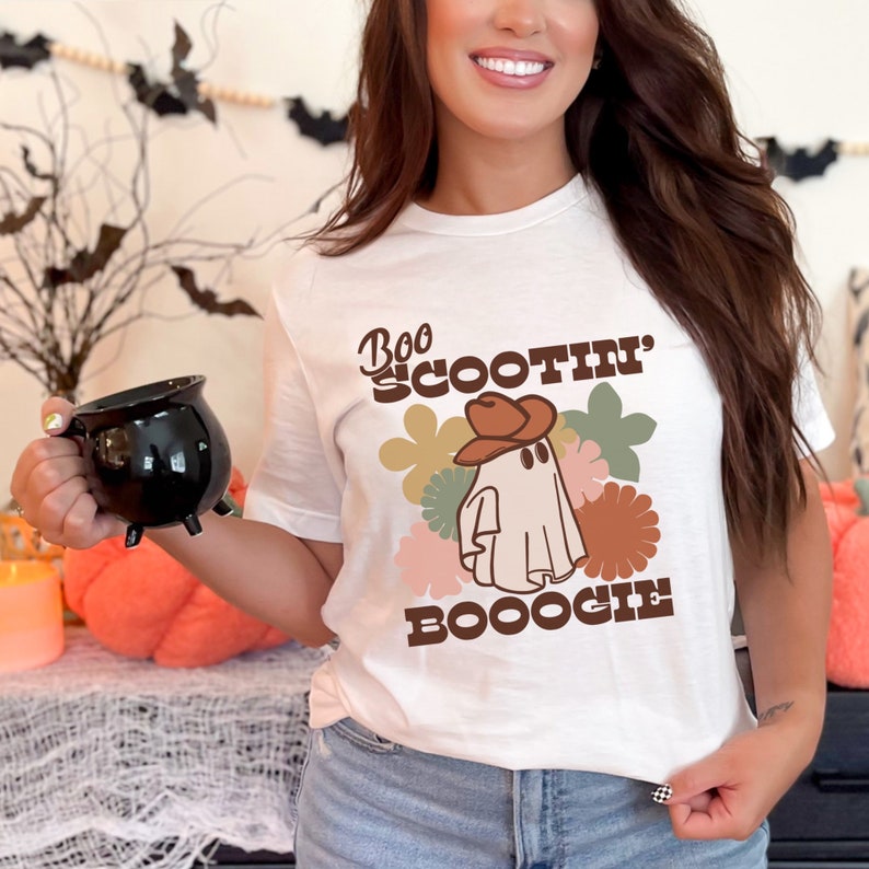 May include: White t-shirt with a brown and white ghost wearing a cowboy hat. The text "Boo Scootin' Booogie" is printed on the shirt.