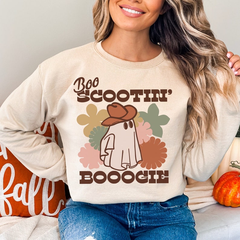May include: A beige sweatshirt with a ghost wearing a cowboy hat and the text "Boo Scootin' Booogie" in brown and white lettering. The ghost is surrounded by colorful flowers.