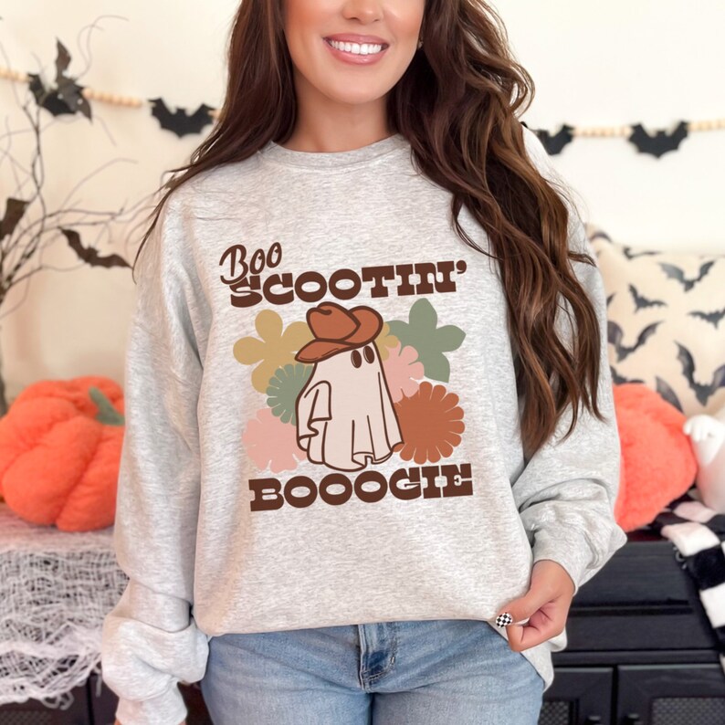 May include: A gray sweatshirt with a white ghost wearing a brown cowboy hat. The ghost is surrounded by colorful flowers. The text "Boo Scootin' Booogie" is printed on the sweatshirt.