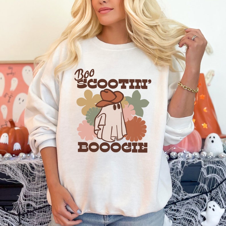 May include: White sweatshirt with a retro-style graphic of a ghost wearing a cowboy hat and the text "Boo Scootin' Booogie".