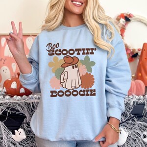 May include: Light blue sweatshirt with a ghost wearing a cowboy hat and the text "Boo Scootin' Booogie" in brown lettering. The ghost is surrounded by colorful flowers.