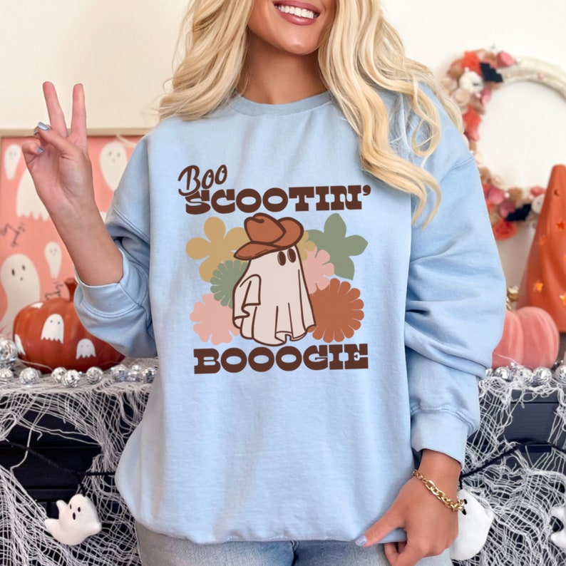 May include: Light blue sweatshirt with a ghost wearing a cowboy hat and the text "Boo Scootin' Booogie" in brown lettering. The ghost is surrounded by colorful flowers.