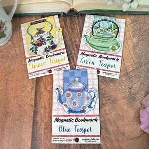 May include: Three magnetic bookmarks featuring teapot illustrations. One has a yellow teapot with floral designs, another a green teapot, and the last a blue teapot with floral accents. Each bookmark has the text "Magnetic Bookmark" and the teapot's name.