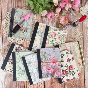 May include: A collection of small notebooks with floral covers, featuring black fabric spines and various rose and botanical patterns. A pink lipstick, a red lipstick, a gold pen, and several gold paper clips are arranged among the notebooks.