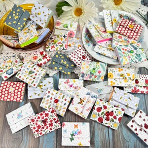 May include: A collection of small, decorative fabric wallets with various floral, butterfly, and insect patterns. The wallets are in different colors, including red, white, and blue, and are arranged on a table with floral accents.