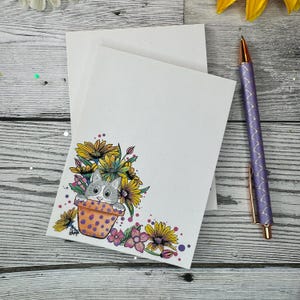 May include: A white notepad with a cute illustration of a cat peeking out of a flower pot with yellow, pink, and purple flowers. The notepad is blank and ready for writing.