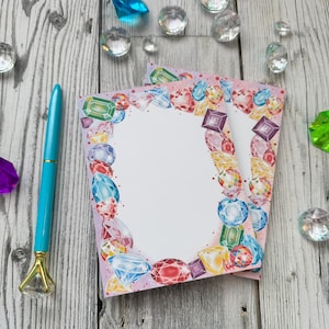 May include: Two blank notepads with a colorful border of gemstones. The border is made up of various shapes and colors of gemstones, including red, blue, green, and purple.