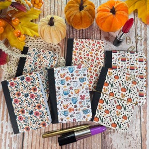 May include: A collection of five small notebooks with black spines and various autumn-themed covers featuring pumpkins, mugs, sweaters, and floral patterns, arranged with two metallic pens and two fabric pumpkins on a light wood surface.