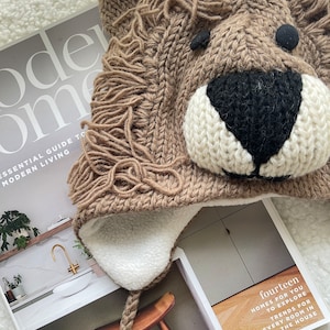 May include: A brown knitted lion hat with a white fleece lining and a long brown cord. The hat has black eyes and a black nose.