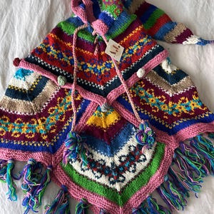 May include: Multicolored knitted poncho with a hood and fringe. The poncho has a geometric pattern and is decorated with pom-poms.