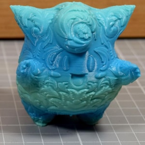 May include: Two decorative figurines, one purple and yellow, the other blue and green, are displayed. Each has an ornate, floral-like design and a smiling face. The figurines are on a grid surface, with a ruler at the bottom.