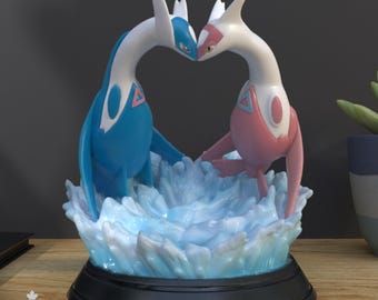 May include: A 3D printed figurine featuring two stylized dragon-like creatures, one blue and one pink, with their heads touching above a translucent blue water-effect base. The figures are mounted on a circular black stand with a flat base.