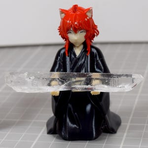 May include: A figurine with bright red hair and cat ears, wearing a black robe, holding a long, clear, jagged quartz crystal. A stack of red, green, yellow, and blue plastic building blocks stands to the left, with a ruler visible at the bottom.