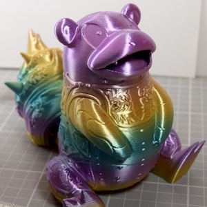 May include: A 3D-printed, rainbow-colored figure of a cartoon-like creature with a large head, small ears, and a wide open mouth. The figure has a detailed design on its chest and back, and is sitting down. The colors transition from purple to yellow.